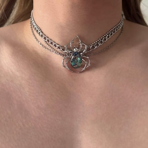 Op de afbeelding: Een zilverkleurige choker-halsketting met meerdere strengen en een centrale spinnenhanger. De spin heeft een frame van gepolijst zilverkleurig metaal en een lichaam met een inleg van iriserende abalone-schelp. De ketting bestaat uit een dikke schakelketting.