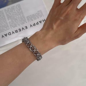May include: A silver-toned chainmail bracelet featuring a repeating pattern of interlocking circular metal rings. The bracelet has a textured, industrial appearance with a matte finish, constructed from multiple small, connected metallic links.