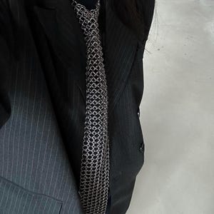 May include: A silver-toned metal chainmail necktie featuring a flexible, interlocking ring construction. The tie is worn over a black shirt and a dark gray pinstriped blazer, showcasing a metallic, textured surface with a uniform pattern.