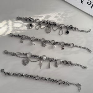 Puede incluir: Cuatro pulseras de cadena en tono plateado con patrones de eslabones intrincados. Las tres pulseras superiores incluyen varios dijes colgantes como piedras negras, perlas blancas, estrellas, soles y formas metálicas, mientras la inferior es una cadena simple.