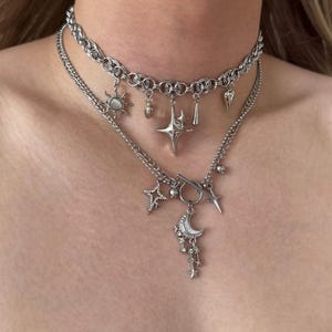 Può includere: Un set di collane a strati color argento composto da un girocollo a catena con ciondoli a forma di sole, stella e goccia. La catena inferiore presenta una chiusura a T, un ciondolo a mezzaluna e ulteriori ciondoli a forma di stella.