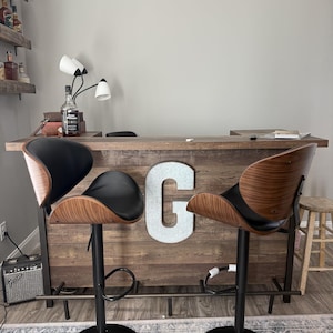 May include: Two modern bar stools with black seats and walnut-colored backs. The stools have black adjustable height bases and are positioned in front of a wooden bar. The bar has a large letter "C" on the front.