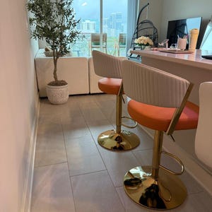 May include: Two bar stools with orange circular seats and white ribbed backrests, supported by gold-coloured metal pedestals with circular bases. The stools are positioned at a counter in a room with light-coloured tile flooring and a tall potted plant.