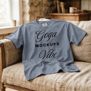 Può includere: Una t-shirt a maniche corte color blu polvere con testo nero stampato sul davanti. Il testo recita Goga MOCKUPS Vibe in una combinazione di caratteri corsivi e stampatello. La maglietta ha un girocollo e un'etichetta del marchio visibile.