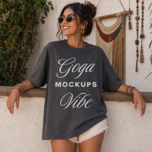 Może przedstawiać: Ciemnoszary t-shirt z krótkim rękawem z białym napisem Goga MOCKUPS Vibe nadrukowanym z przodu. Koszulka ma okrągły dekolt i luźny krój, a słowa Goga i Vibe są zapisane kursywą, natomiast MOCKUPS drukowanymi literami.