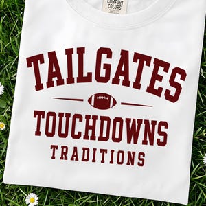 Może przedstawiać: Biały t-shirt z krótkim rękawem i okrągłym dekoltem, ozdobiony bordowym napisem TAILGATES, TOUCHDOWNS i TRADITIONS, z małą grafiką piłki do futbolu amerykańskiego umieszczoną między dwiema pierwszymi liniami oraz metką marki Comfort Colors.