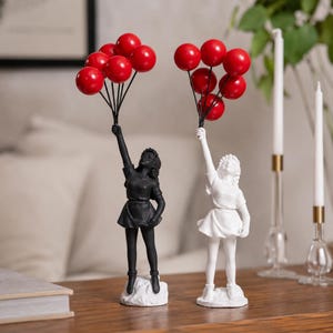 Puede incluir: Dos figuras decorativas de mujeres sosteniendo globos rojos. Una figura es negra, la otra blanca. Cada figura lleva un vestido de manga corta y se apoya en una pequeña base. Los globos son de color rojo vivo.