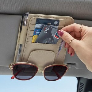 Puede incluir: Un organizador de visera para coche en color beige con múltiples ranuras para tarjetas de crédito, un lazo para un bolígrafo y un clip para gafas de sol. El organizador está sujeto a la visera y contiene varias tarjetas y un bolígrafo.