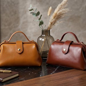 May include: Two structured handbags with a top handle and a central snap closure. One bag is a mustard yellow color, and the other is a deep reddish-brown. Both feature a smooth, rounded frame design with a single strap and gold-toned hardware.