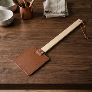 May include: A fly swatter featuring a rectangular brown leather head with perforated holes, attached to a light-coloured wooden handle with two metal rivets. A thin brown leather cord is tied through a hole at the end of the wooden handle.