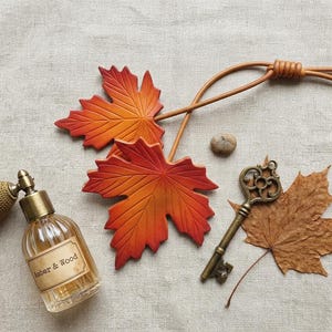 May include: A glass perfume bottle with a gold atomizer bulb and a label reading Amber & Wood, two orange and red maple leaf ornaments on a leather cord, a small brown stone, an antique brass key, and a dried brown maple leaf.