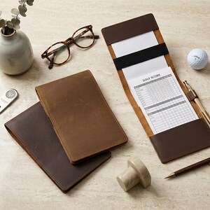 May include: Two brown leather golf scorecard holders, one closed and one open with a white golf score card inserted. Accessories include a silver metal divot tool, a white golf ball, a gold pen, a brown pen, and tortoise shell glasses.