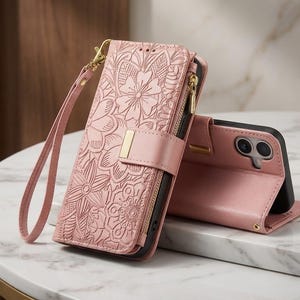 May include: A dusty pink wallet-style phone case featuring an embossed floral pattern, a gold-toned metal accent on the magnetic closure, a side zip compartment, and a detachable wrist strap. The case is shown in both closed and open positions.