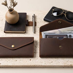 May include: Two dark brown leather envelope-style wallets with brass snap closures. One wallet is closed, while the other is open, revealing paper currency and a smartphone inside. A small leather notebook, a metal pen, and sunglasses are nearby.