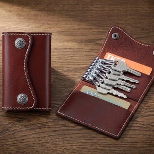 May include: A dark brown leather key case with white stitching and two silver-toned metal snap buttons featuring embossed circular patterns. The open case reveals a metal key holder with six silver keys and two interior card slots.