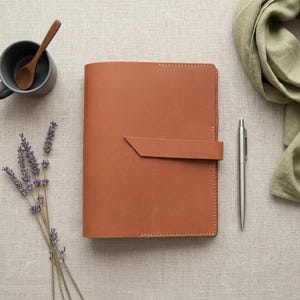 May include: A tan leather notebook with a horizontal strap closure, positioned next to a silver metal pen, a dark gray ceramic mug with a wooden spoon, a sprig of dried lavender, and a folded sage green fabric scarf on a neutral linen surface.