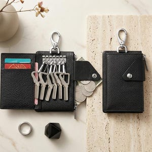 May include: Two black leather key cases with silver hardware. One case is open, revealing six metal key hooks, card slots with colorful cards, and a zippered compartment containing coins. The other case is closed with a snap button closure.