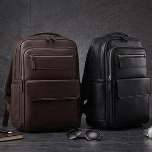 May include: Two backpacks, one in dark brown and one in black, each featuring a top handle, a zippered main compartment, a front zippered pocket, and a lower flap pocket with silver-toned hardware, presented side by side in a clean arrangement.