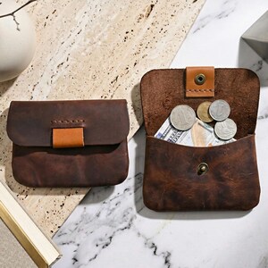 May include: Two dark brown leather pouches with a smooth matte finish. One pouch is closed with a tan leather tab, while the other is open, revealing a metal snap button closure, several silver-colored coins, and a folded paper bill inside.