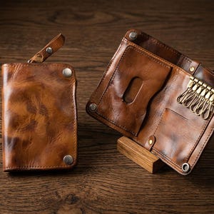 May include: A brown leather key case wallet featuring a smooth, distressed finish, silver-toned metal snap closures, and a brass-colored six-hook key organizer. The interior includes a card slot with a cutout and a leather pull tab on the exterior.