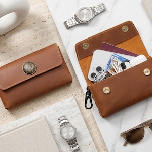 May include: Two brown leather pouches with snap closures and black carabiner clips. One pouch is closed and features a circular metal emblem on the flap. The other pouch is open, revealing a smartphone, keys, a passport, and white earbuds inside.