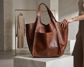 May include: A large, slouchy brown leather tote bag with a smooth, textured finish. The bag features a wide, open-top design, two integrated shoulder straps, and a prominent, deep exterior pocket on the front panel with visible stitching details.