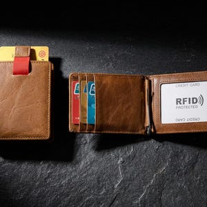 May include: A tan leather bifold wallet with a red pull tab and a metal money clip. The interior features multiple card slots and a clear ID window with a white card printed with the text RFID PROTECTED and CREDIT CARD.