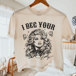 Könnte beinhalten: Ein elfenbeinfarbenes Kurzarm-T-Shirt mit einem schwarzen Grafikdruck einer Frau mit voluminösem Haar, Schmetterlingen und Sternen. Der Text "I BEG YOUR" ist in einer fetten, geschwungenen Schrift darüber gedruckt, mit dem Namen "Dolly" in Schreibschrift.
