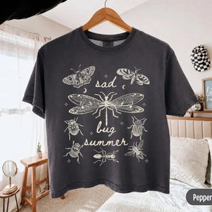 Pode incluir: Uma t-shirt de manga curta cinzento-carvão com uma impressão gráfica em cor creme de vários insetos, incluindo uma libélula, traças e escaravelhos, com o texto "sad bug summer" centrado entre as ilustrações em letra cursiva.