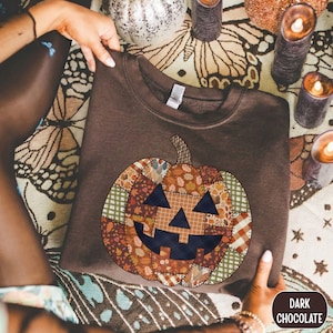 May include: A dark chocolate brown crewneck sweatshirt featuring a large central graphic of a jack-o'-lantern pumpkin. The pumpkin is designed with a patchwork pattern of various autumn-themed fabrics, including plaid, gingham, and leaf prints.