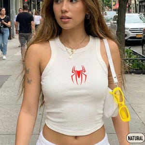 Pode incluir: Uma regata branca sem mangas com um gr&aacute;fico de aranha vermelha centralizado no peito. A pe&ccedil;a tem uma gola redonda alta e uma silhueta justa. Uma bolsa de ombro branca com al&ccedil;a est&aacute; presente e &oacute;culos amarelos est&atilde;o fixados nela.