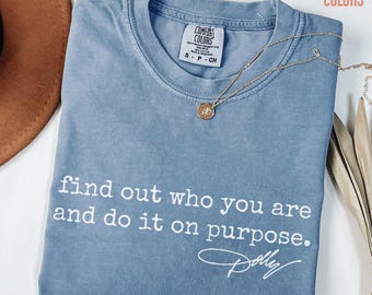 May include: A blue jean coloured t-shirt with a crew neckline and a Comfort Colors brand label. The shirt features the white printed text: find out who you are and do it on purpose. Dolly. A gold chain necklace with a small round pendant rests on the fabric.