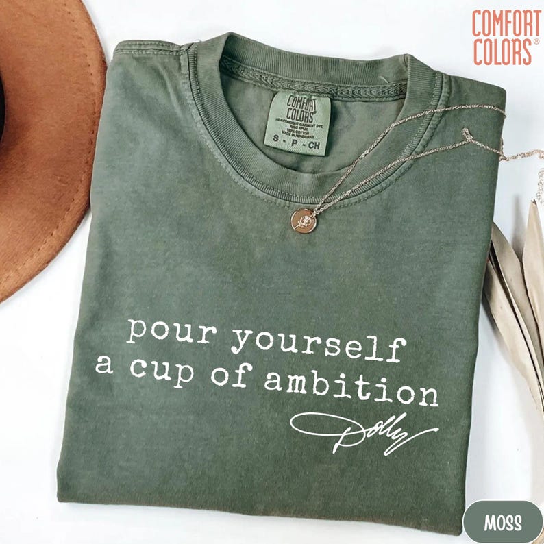 Puede incluir: Una camiseta de cuello redondo color verde musgo con el texto en blanco "pour yourself a cup of ambition" seguido de una firma que dice "Dolly". La camiseta tiene una etiqueta de la marca Comfort Colors en el cuello y una fina cadena dorada.