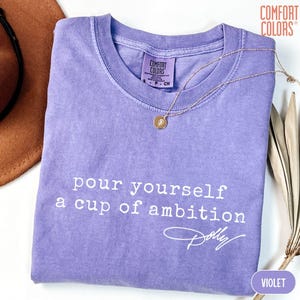 Puede incluir: Una camiseta violeta de manga corta con cuello redondo y una etiqueta de la marca Comfort Colors. La prenda presenta un texto en blanco que dice "pour yourself a cup of ambition" seguido de una firma "Dolly" en letra cursiva.