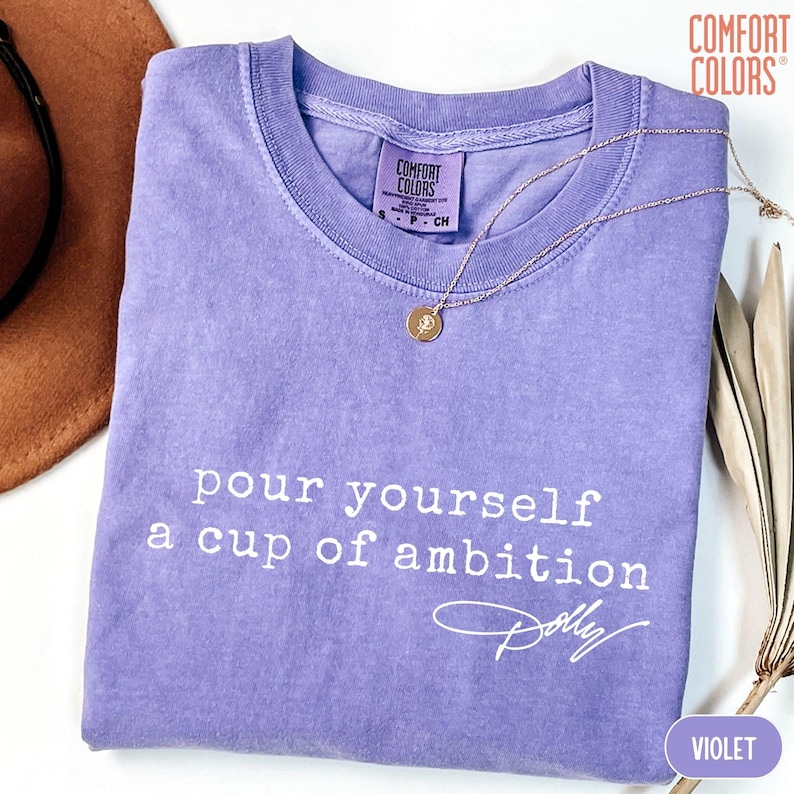 Puede incluir: Una camiseta violeta de manga corta con cuello redondo y una etiqueta de la marca Comfort Colors. La prenda presenta un texto en blanco que dice "pour yourself a cup of ambition" seguido de una firma "Dolly" en letra cursiva.
