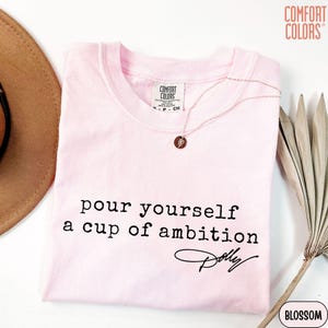 Puede incluir: Una camiseta de manga corta de color rosa claro con el texto impreso en negro "pour yourself a cup of ambition" seguido de la firma "Dolly". La camiseta tiene una etiqueta de la marca Comfort Colors en el cuello y un collar dorado fino.