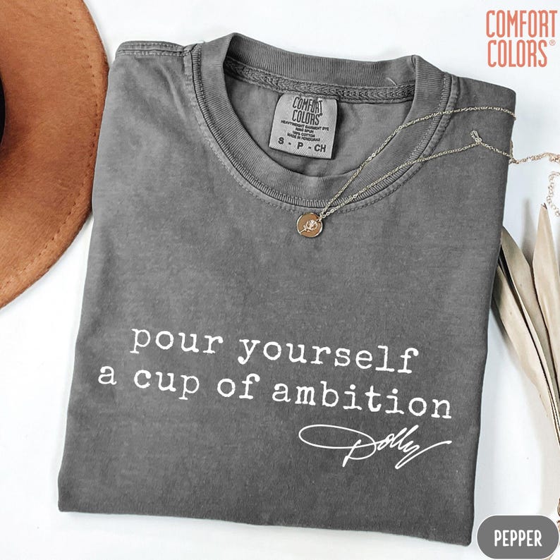 Puede incluir: Una camiseta de manga corta gris carb&oacute;n con el texto blanco "pour yourself a cup of ambition" y una firma debajo. La camiseta presenta una etiqueta de la marca Comfort Colors en el cuello interior y un collar de cadena dorada.