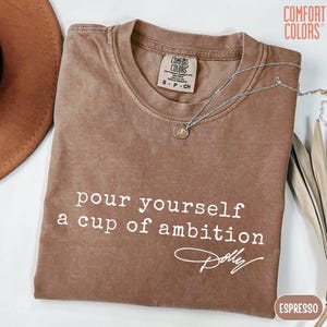 Puede incluir: Una camiseta marr&oacute;n de manga corta con el texto blanco "pour yourself a cup of ambition" y una firma debajo. La prenda incluye una etiqueta de la marca Comfort Colors en el cuello interior y un collar de cadena plateada sobre la tela.