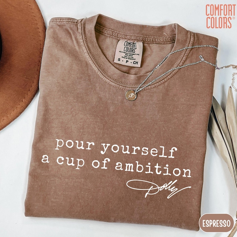 Puede incluir: Una camiseta marr&oacute;n de manga corta con el texto blanco "pour yourself a cup of ambition" y una firma debajo. La prenda incluye una etiqueta de la marca Comfort Colors en el cuello interior y un collar de cadena plateada sobre la tela.