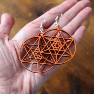 May include: A pair of orange wooden earrings with a geometric star design. The earrings are made of wood and have a silver hook.