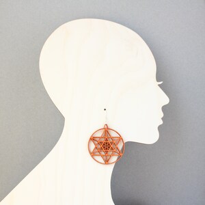 May include: A pair of wooden earrings with a geometric star design. The earrings are a reddish-brown colour and are hanging from a mannequin's ear.