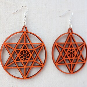May include: A pair of orange wooden earrings with a geometric star design. The earrings are made of wood and have a silver hook.