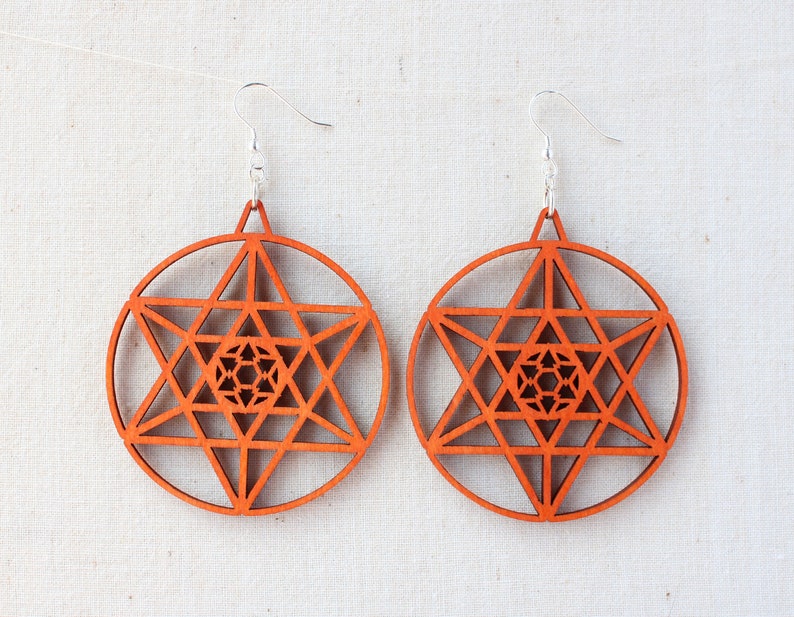 May include: A pair of wooden earrings with a geometric star design. The earrings are orange and have a silver hook.