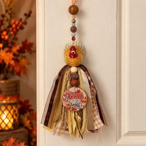 May include: A hanging decoration featuring a turkey figurine atop a tassel made of brown, gold, and white ribbons and fabric strips. A round wooden tag hangs from the center with the text GOBBLE TIL YOU WOBBLE and autumn imagery.