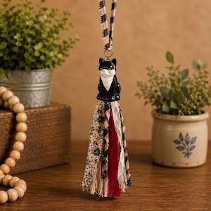 May include: A hanging ornament featuring a black and white cat figurine attached to a multi-textured fabric tassel. The tassel consists of strips of patterned fabric in beige, black, and deep red, suspended by a black and beige checkered ribbon.