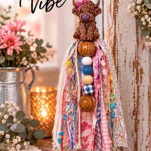 May include: A hanging ornament featuring a brown highland cow figurine with a pink bow, suspended above a tassel made of assorted fabric strips in pink, white, and blue patterns, accented with wooden and blue gingham beads and a heart charm.