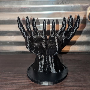 May include: A black 3D printed sculpture of two hands reaching upwards, with long, slender fingers. The sculpture is mounted on a circular base.