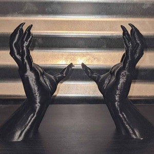 May include: A pair of black 3D printed hands with long fingers, reaching up towards the light. The hands are posed as if they are holding something.