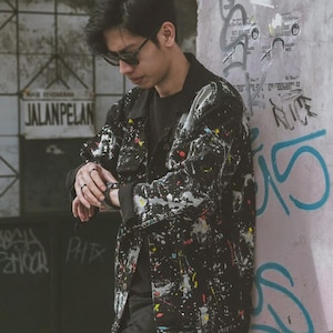May include: A black denim jacket featuring a multi-colored paint splatter pattern in white, yellow, red, and blue. The jacket has a classic collar, long sleeves, and a button-front closure, worn over a plain black shirt with black trousers.