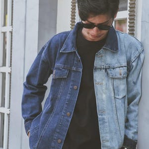 May include: A two-tone denim jacket featuring a dark blue wash on the right side and a light blue wash on the left side. The jacket includes a classic collar, metal button closures down the front, two chest pockets with button flaps, and long sleeves.
